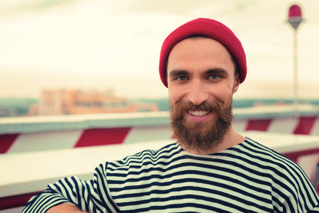 Attractive man. Handsome bearded young man looking happy and smiling while standing in his striped clothes and red hatの写真素材