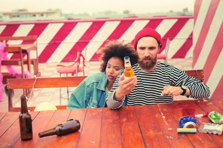 Looking at beer. Tired young man sitting at the table with sleepy girlfriend and looking at the bottle of beer after partyの写真素材