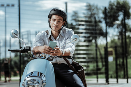 Group chat. Handsome male student using phone and sitting on motorbikeの写真素材