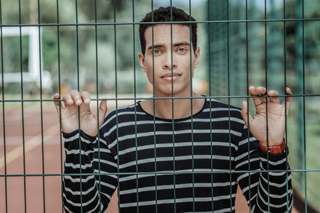 Join me. Appealing sincere guy staying behind fence and gazing at camera