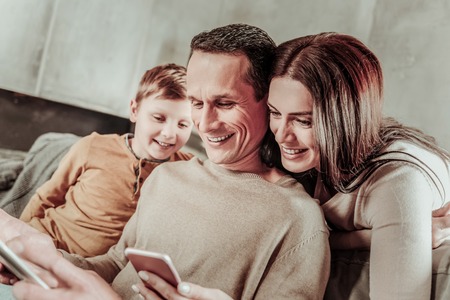 Warm atmosphere. Cheerful boy sitting near his daddy while looking at screen of gadget