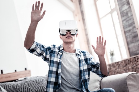 Virtual reality. Calm young man sitting on the sofa and putting his hands up while wearing virtual reality glassesの写真素材