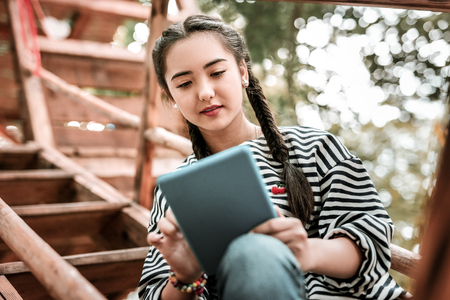 Active messaging. Cheerful Asian female keeping smile on her face while looking at screen of tablet