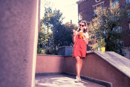Cute girl. Cute funny appealing girl wearing bright red sunglasses and white earphones drinking some refreshing drinkの写真素材