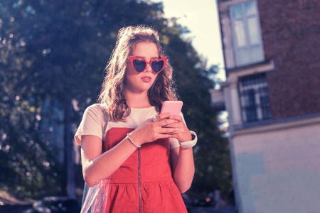 Texting boyfriend. Dark-haired fashionable student wearing bright red sunglasses holding her pink phone while texting boyfriendの写真素材