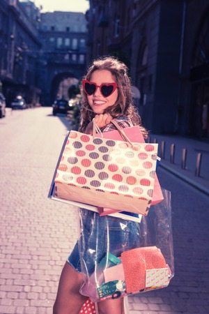 Cheerful shopaholic. Cheerful dark-haired shopaholic feeling very satisfied while leaving boutique with many shopping bagsの写真素材
