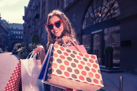 Joyful shopping. Dark-haired curly woman wearing red sunglasses feeling extremely cheerful while enjoying shopping timeの写真素材