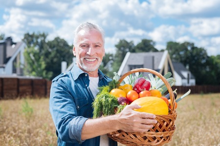 Peppers and tomatoes. Dark-eyed bearded farmer smiling broadly while holding basket full of red sweet peppers and tomatoesの写真素材