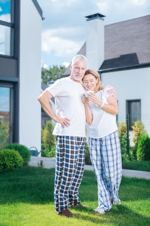Coffee outside. Cute loving couple wearing baggy pajama trousers and white shirts drinking coffee outside the houseの写真素材