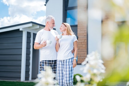 Coffee in courtyard. Bearded caring man hugging his happy beautiful wife while drinking coffee in their courtyardの写真素材