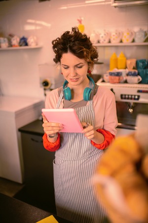 Professional barista. Cheerful curly haired female holding gadget in both hands while enjoying good newsの写真素材