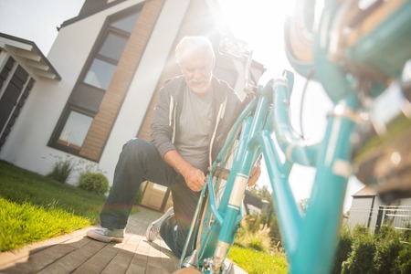 Modern man. Modern elderly grey-haired man wearing blue sneakers repairing his old blue bicycleの写真素材