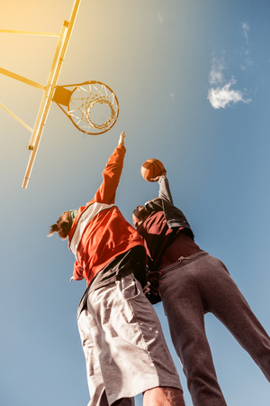 Hard game. Low angle of young well built men standing under the basketball basket while throwing the ballの写真素材