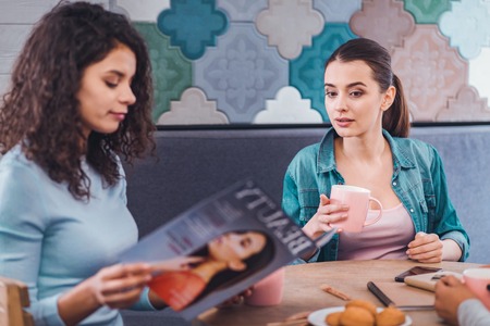What are you reading. Nice beautiful woman asking a question to her friend while sitting with her at the tabletの写真素材