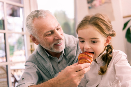 Fresh croissant. Cute dark-eyed granddaughter biting just baked fresh croissant sitting near her grandfatherの写真素材