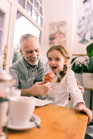 Stylish granddaughter. Cute stylish granddaughter wearing white shirt trying croissant having breakfast with grandfatherの写真素材