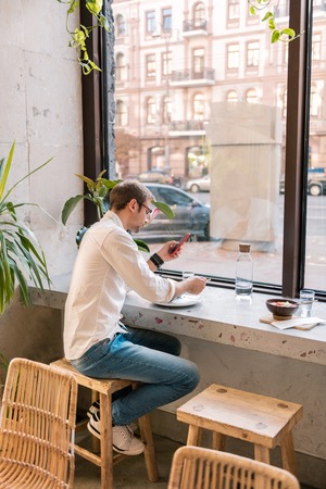 Man in cafe. Stylish handsome dark-haired man wearing jeans and white shirt sitting in atmospheric vegan cafeの写真素材