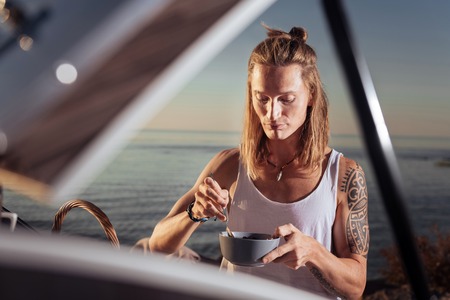Cereals in morning. Man with tattoo living in compact trailer standing in nature eating cereals in morningの写真素材