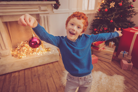 Cheerful mood. Upbeat red-haired preteen boy spreading his arms wide and posing for the camera while holding a Christmas ball in his handの写真素材