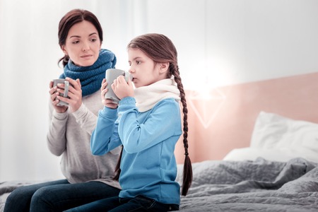 Home atmosphere. Portrait of pleasant family sitting on the bed while holding cups and drinking tasty teaの写真素材
