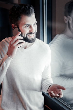 Where are you. Cheerful young male keeping smile on his face and looking at window while talking per telephoneの写真素材