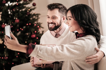 Doing selfie. Cheerful bearded man expressing positivity while embracing his girlfriend and posing on cameraの写真素材