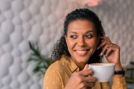 Cup of coffee. Portrait of beautiful dark-haired curly woman wearing yellow blouse enjoying her cup of coffeeの写真素材