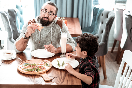 Busy boy. Cute dark-eyed boy wearing squared shirt feeling busy while chewing his piece of pizzaの写真素材