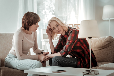 Serious conversation. Short-haired distracted woman cheering up her upset friend sitting on the couch in living roomの写真素材