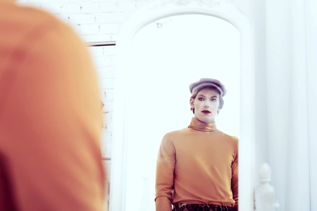Transgender sad man. Transsexual man looking in his reflection in the mirror being fully-dressed and with make up onの写真素材