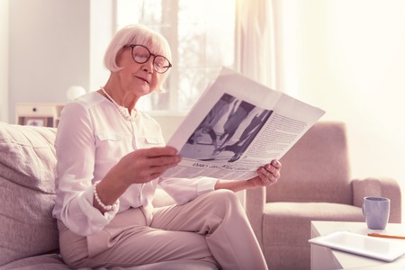 Curious old lady. Curious old lady attentively reading newspaper wearing big clear glasses and light clothesの写真素材