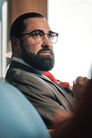 Economist in glasses. Dark-haired bearded economist wearing glasses sitting on sofa in his hotel roomの写真素材
