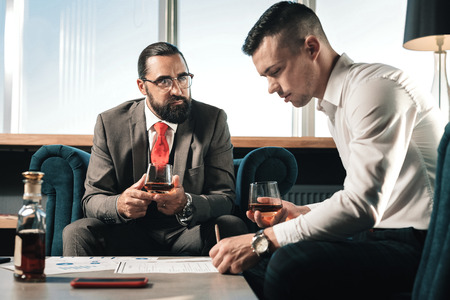 Working and drinking. Bearded boss watching his young trainee working and drinking whisky