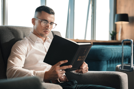 Holding Bible. Dark-haired man wearing glasses holding the Holy Bible enjoying reading time