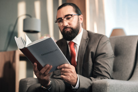 Reading about law. Experienced bearded lawyer reading administrative law while sitting in the officeの写真素材