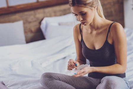 For convenience. Beautiful young woman sitting on the bed and putting a blue capsule into pill organizer, rearranging daily dosesの写真素材