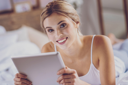 Close up of beaPeaceful day. The close up of a gorgeous young woman smiling at the camera while lying on the bed and using her tablet utiful young woman using tablet on bedの写真素材