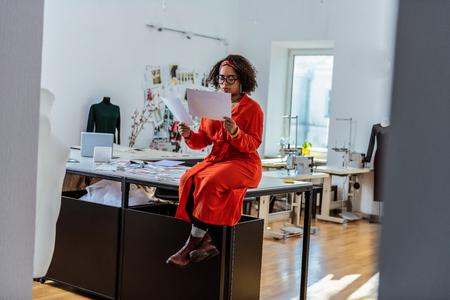 Looking for inspiration. Bright interesting young woman crossing her legs while sitting on working table in studioの写真素材