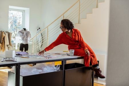 Observing photo materials. Short-haired mixed face female reaching out one of the photos on the table top while sitting on the cornerの写真素材