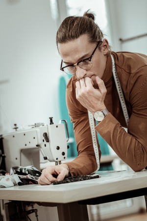 Right choice. Young tall dressmaker in eyeglasses looking involved while thinking about fabric for a...