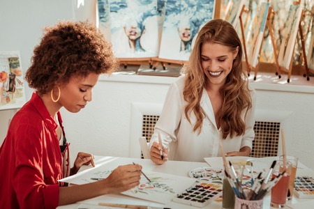 Broad smile. Beaming blonde-haired artist wearing white blouse smiling broadly painting with friendの写真素材