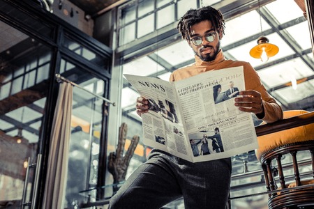 News review. Delighted international male wearing glasses while staring at newspaperの写真素材