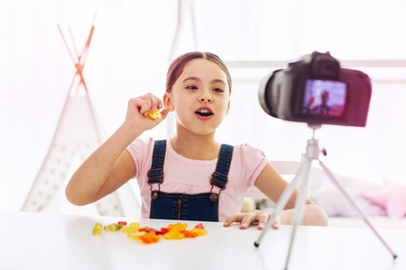 Girl eating. Funny dark-eyed girl wearing jeans jumpsuit sitting in front of camera eating gummy bearsの写真素材