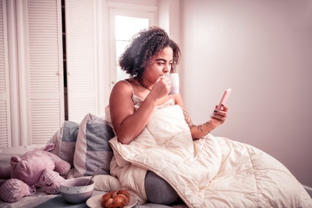Coffee and breakfast. Attentive short-haired woman texting messages on her smartphone while drinking morning teaの写真素材