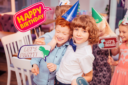 Making fun. Handsome curly-haired boy embracing his friend while having fun at partyの写真素材