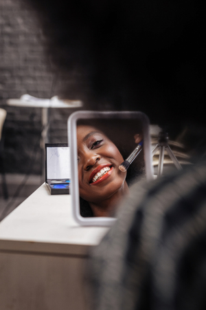 Beautiful smile. Cute dark-skinned woman with coral lipstick smiling positively while doing makeupの写真素材
