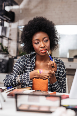 Lip gloss. Amazing african american woman with curly hair looking concentrated while trying on new lip glossの写真素材