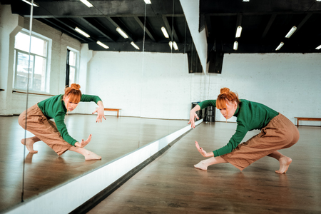 Modern dance. Pretty red-haired professional dancer wearing orange pants dancing near the mirrorの写真素材