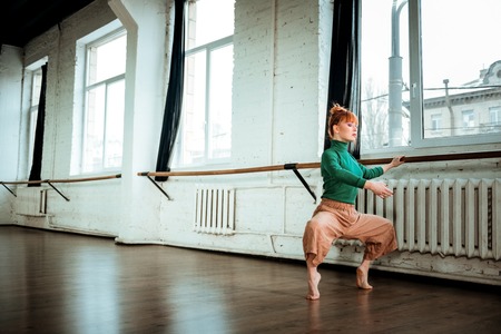 Sportive woman. Young professional modern dancer with red hair looking involved while training near ballet barの写真素材