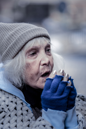 Keeping warm. Depressed aged woman breathing at her hands while trying to keep warmの写真素材